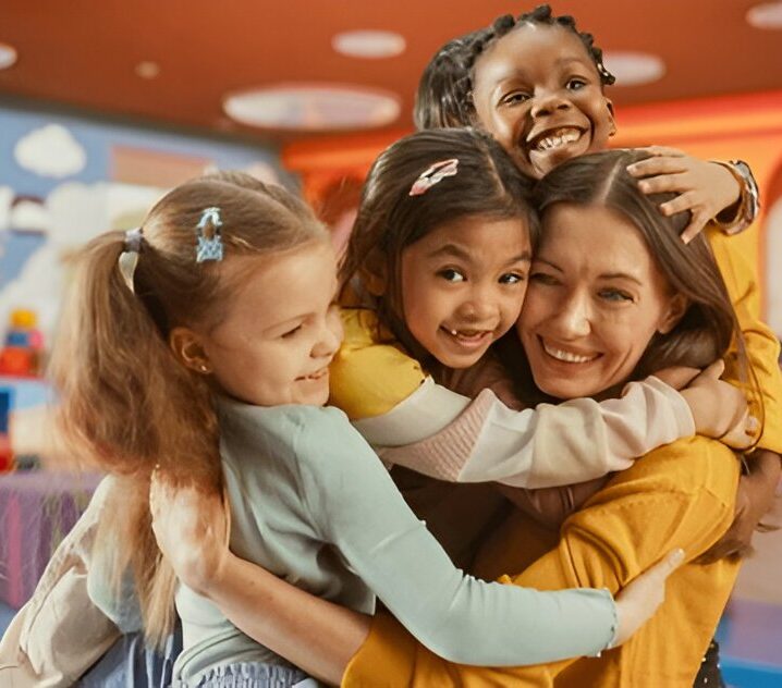 Children hugging a smiling female caregiver in a colorful daycare setting.
